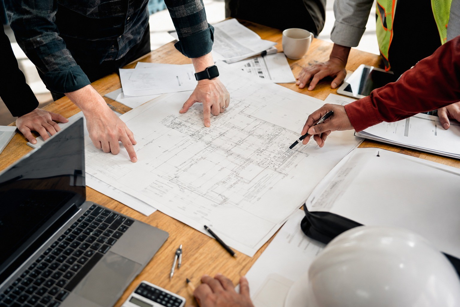 A team reviewing construction drawings around a table with a laptop, hard hat, and calculator